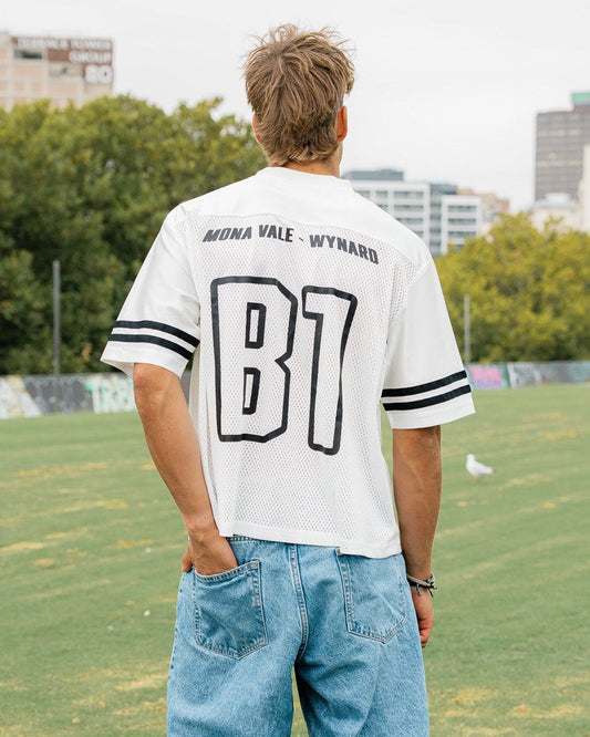 Person wearing a white sports jersey with 'B1' on the back, standing on a grassy field with trees and buildings in the background.