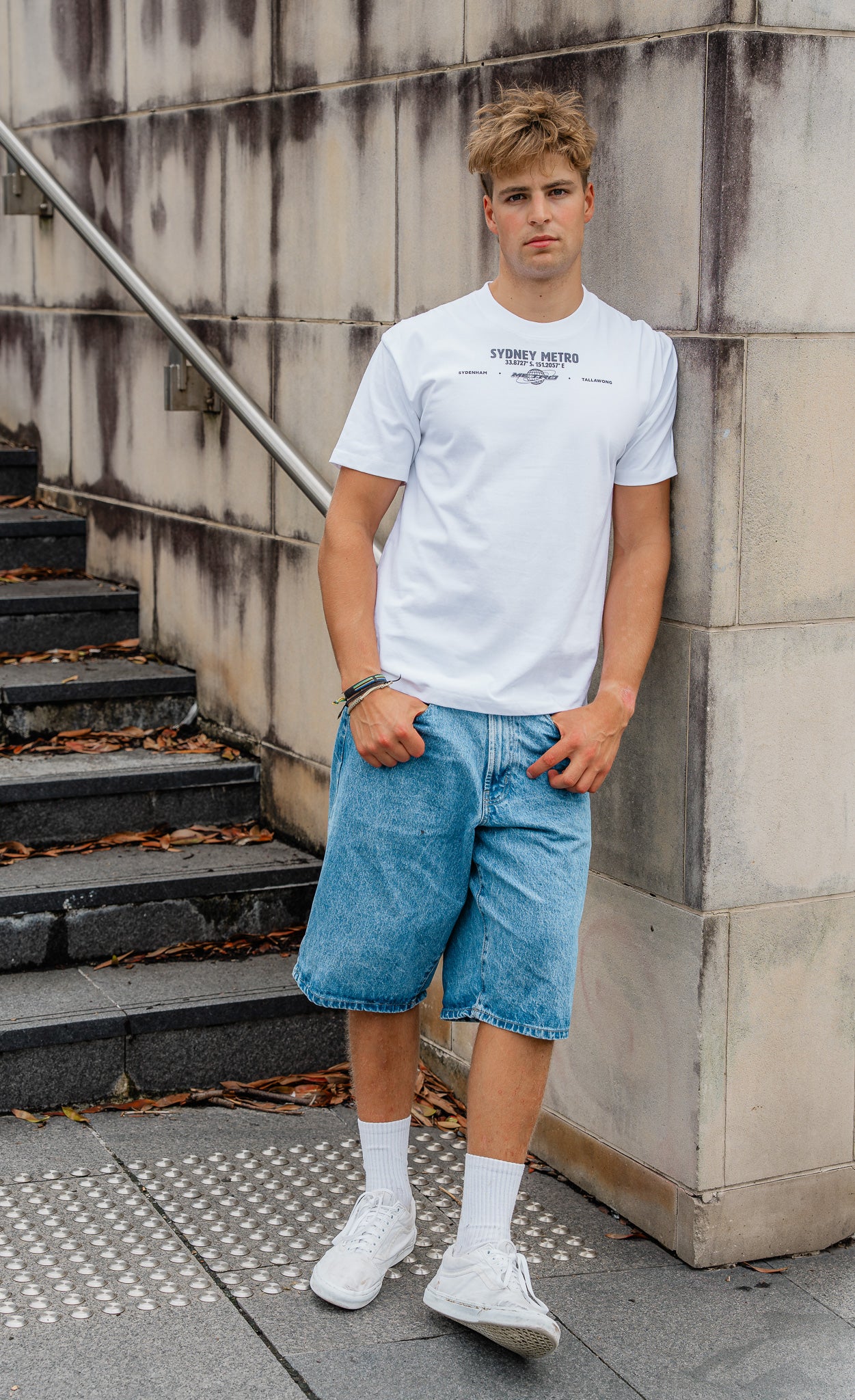 Man wearing a white t-shirt and blue shorts standing against a concrete wall.