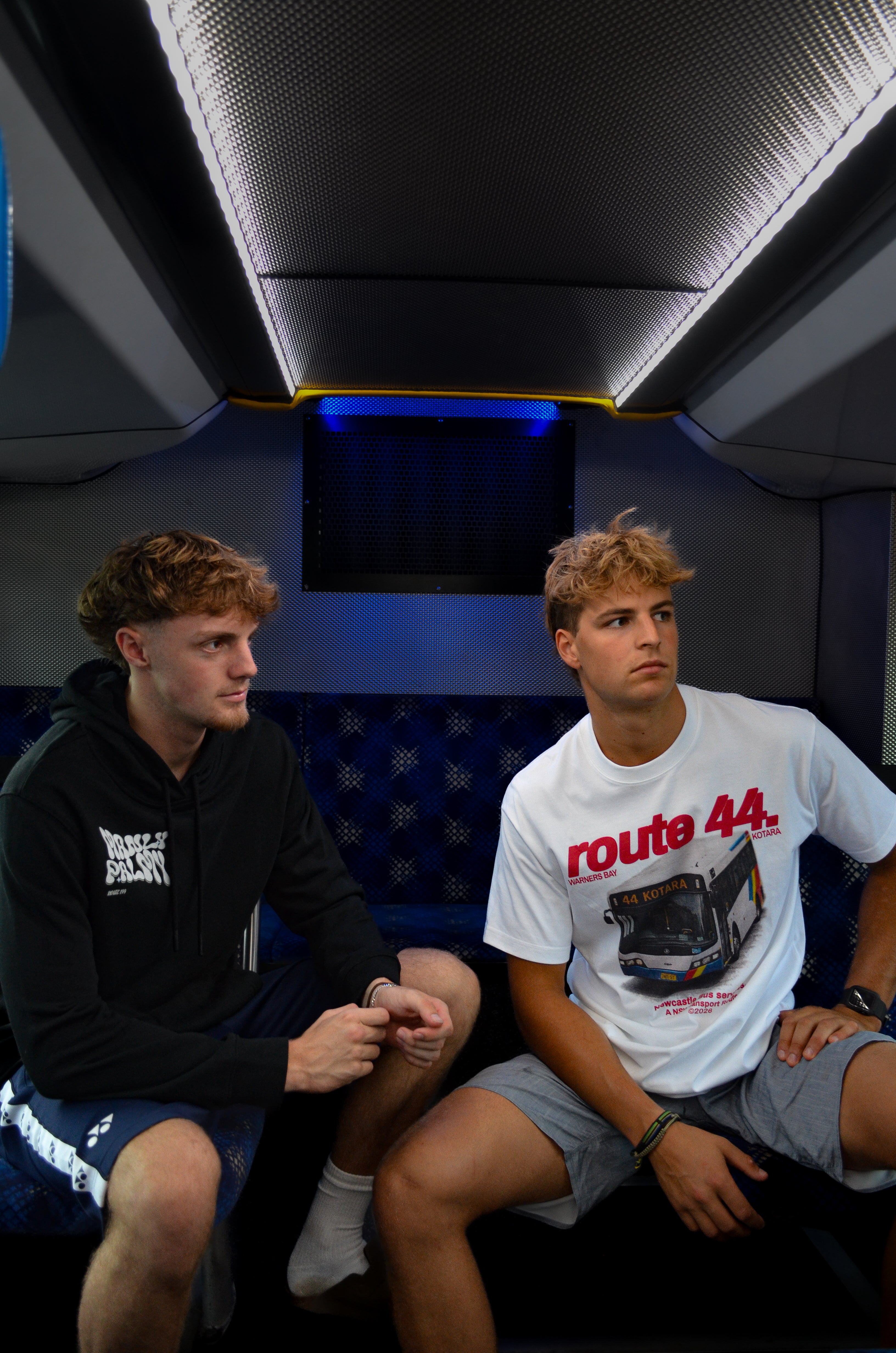 Two young men sitting inside a vehicle, one wearing a black hoodie and the other in a white t-shirt with 'route 44' design.