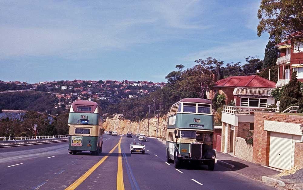 Two vintage double-decker buses on a road with houses and hills in the background.