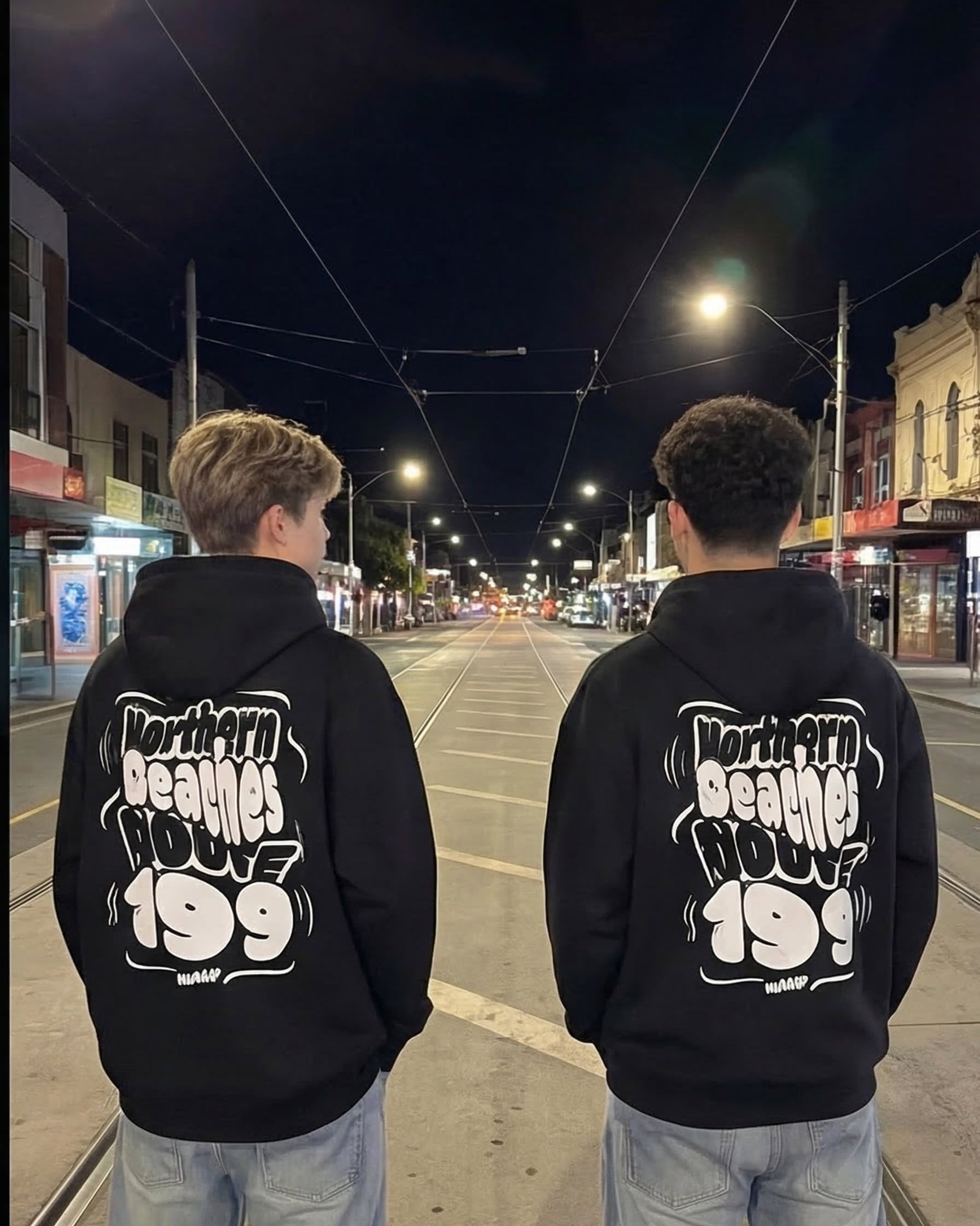 Two people wearing black hoodies with 'Northern Beaches House 199' text on a street at night.
