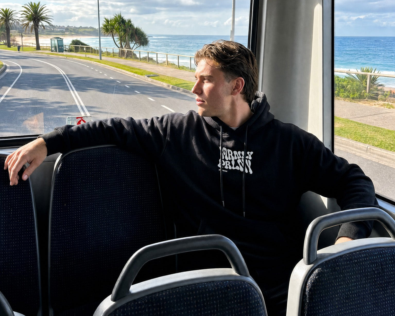 A man wearing a black hoodie with a white graphic sits inside a moving bus, looking out the window at a coastal road and ocean view.